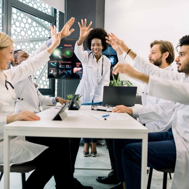 Grupo de doutores ou cientistas motivados, sentado ao redor da mesa na sala de conferências da clínica moderna, dando um gesto de cinco com as mãos, depois de um ótimo trabalho em equipe.